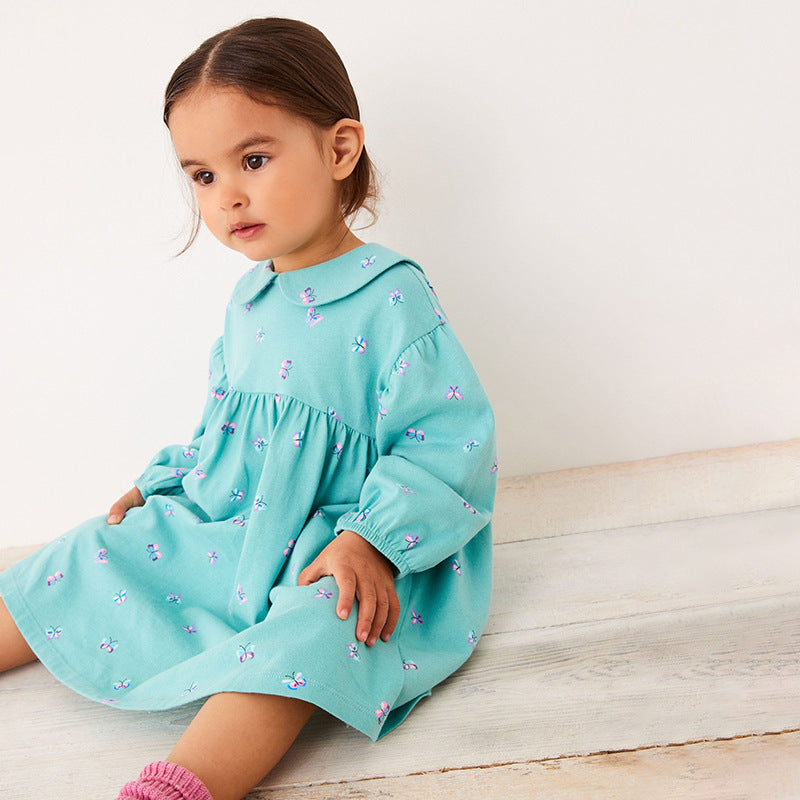 Young girl wearing a light blue dress with floral patterns sitting on a wooden floor.