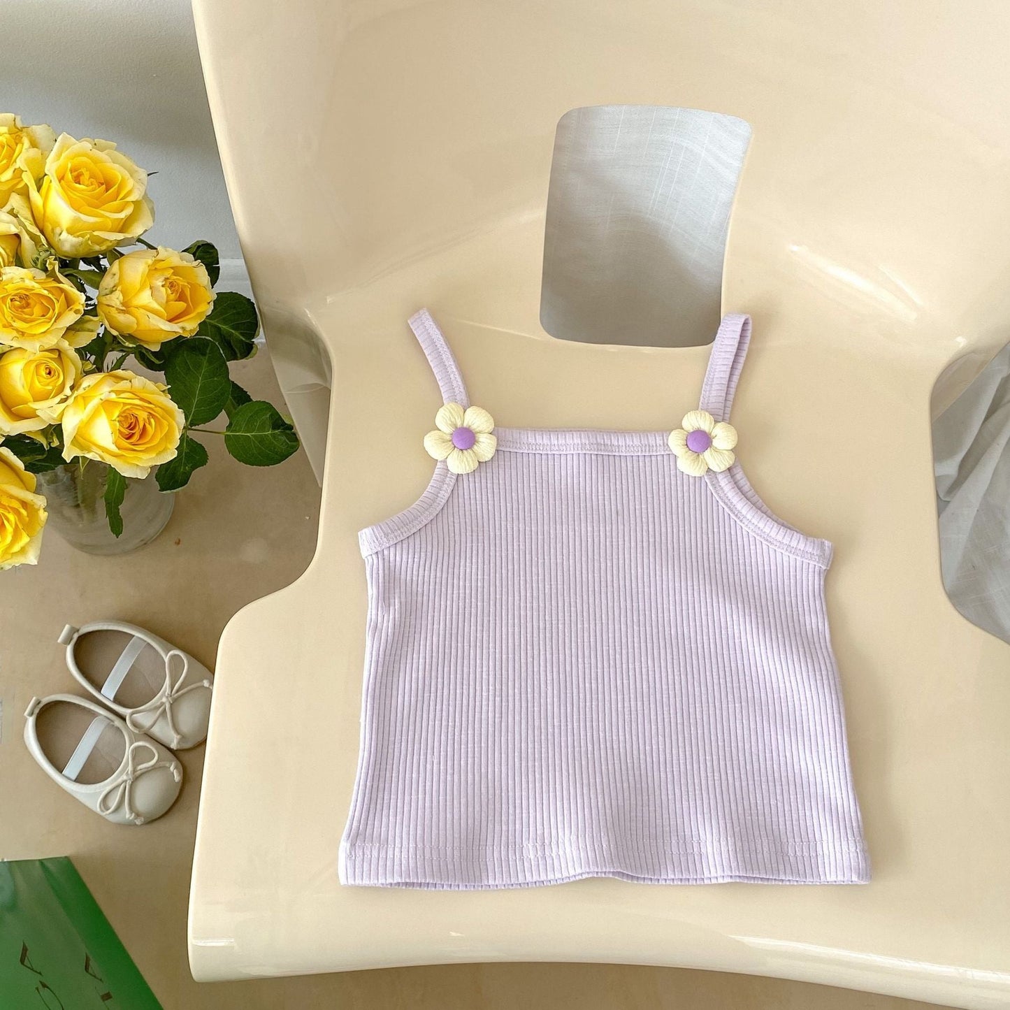 Purple ribbed tank top with floral straps on a white chair next to yellow roses and baby shoes.