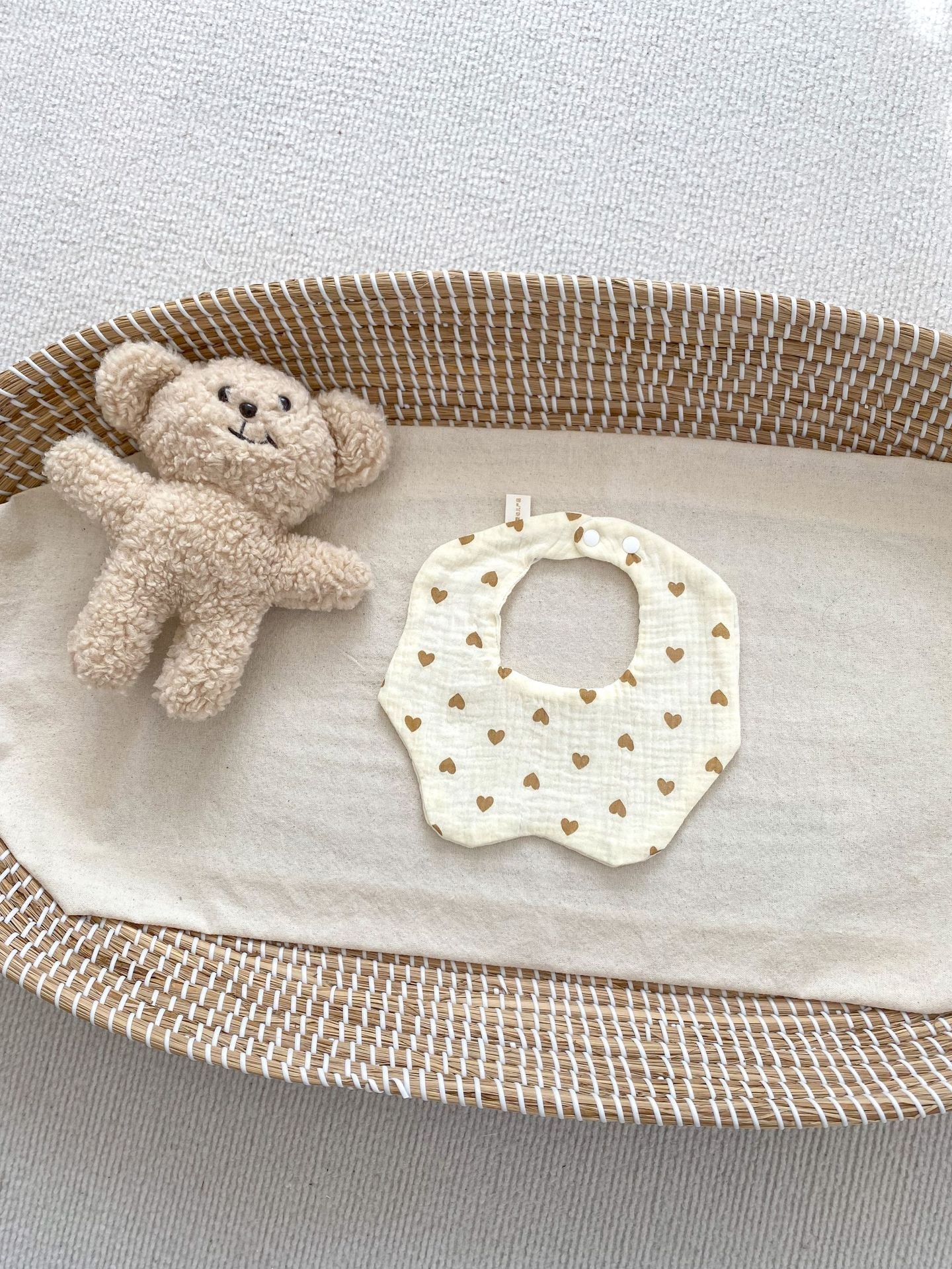 Baby bib with heart pattern and teddy bear in a woven basket on a light background