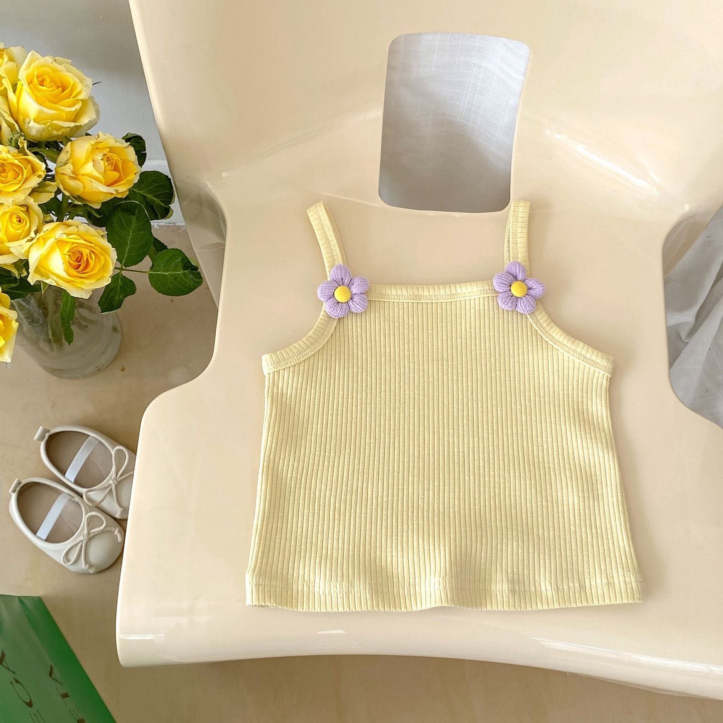 Yellow ribbed tank top with floral straps on a mannequin, next to yellow roses and baby shoes.
