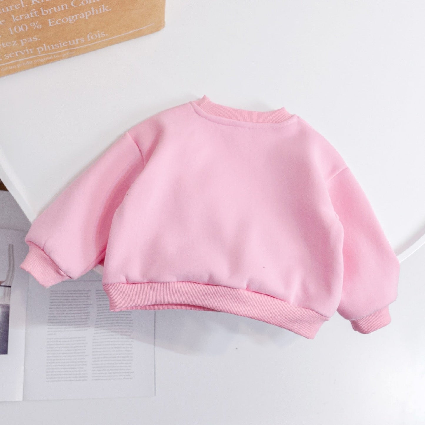 Pink children's sweatshirt on a white surface with a cardboard box in the background
