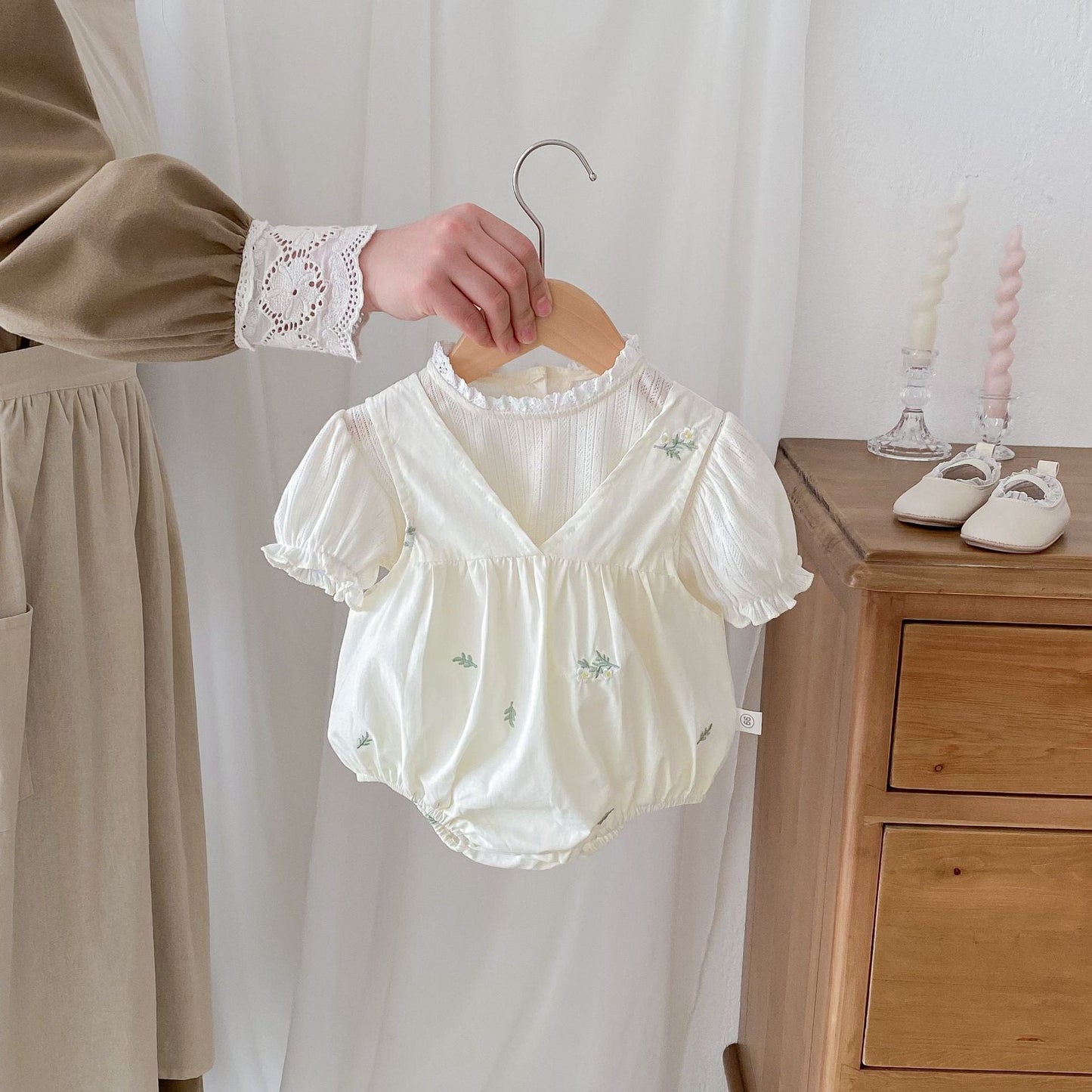 Baby romper with floral embroidery on a hanger, held by a person in a neutral setting.