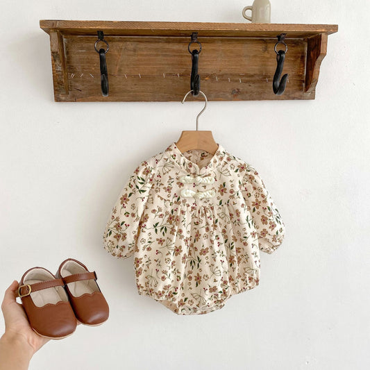 Floral baby romper on a hanger with brown shoes held in front, against a wooden shelf background.