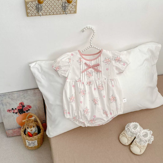 Baby outfit with floral design on a hanger, next to baby shoes and a teddy bear in a basket.