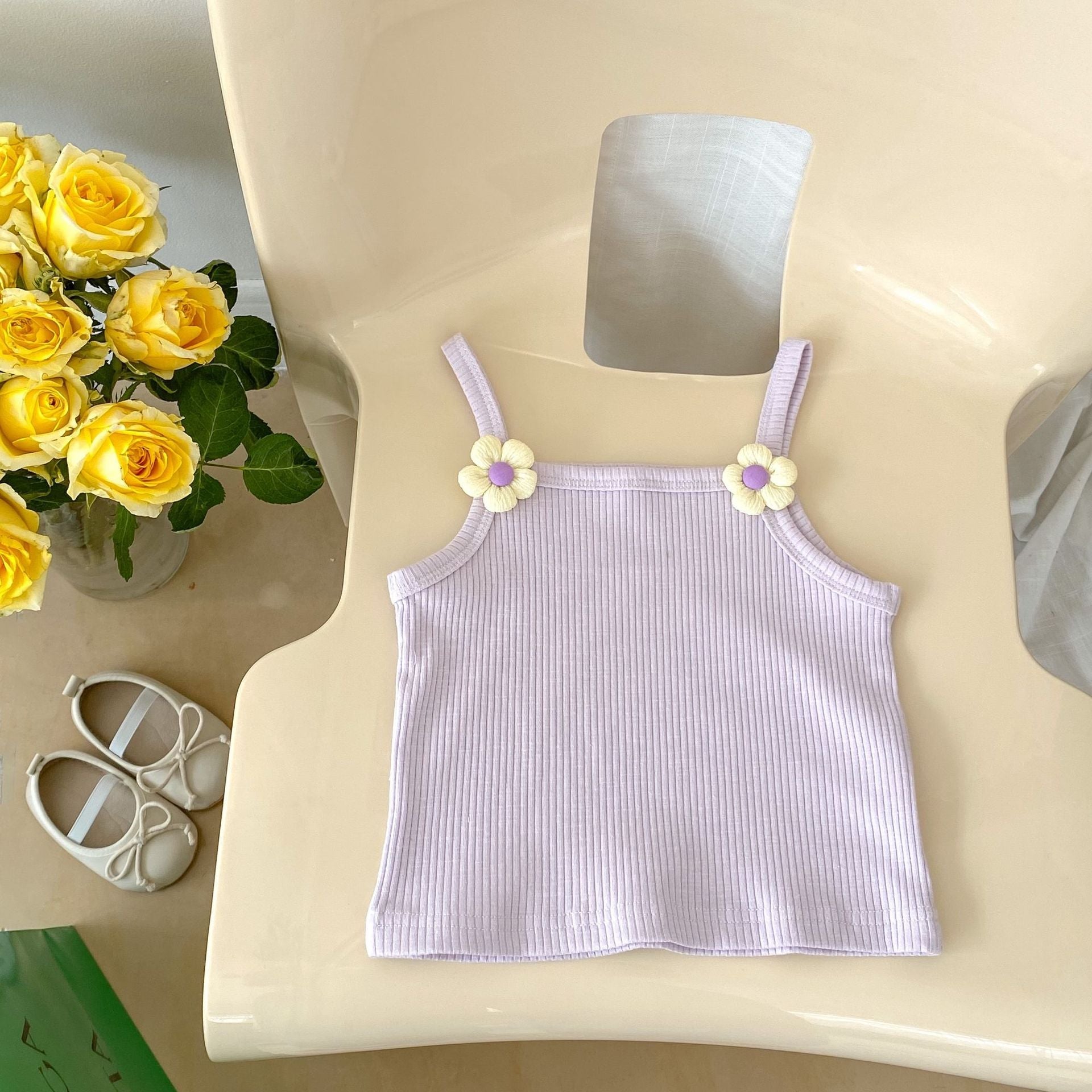 Purple ribbed tank top with floral straps on a white chair next to yellow roses and baby shoes.