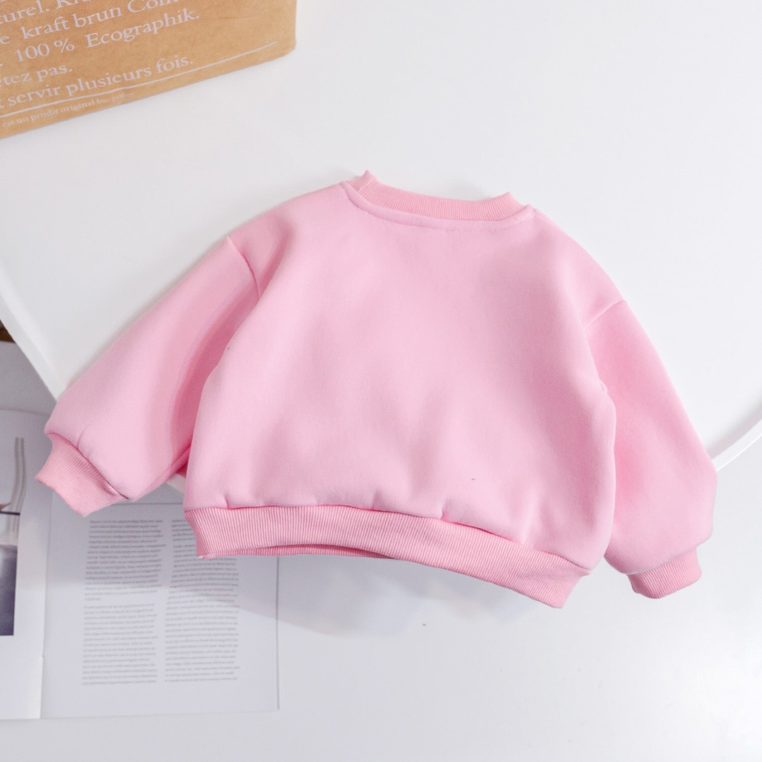 Pink children's sweatshirt on a white surface with a cardboard box in the background