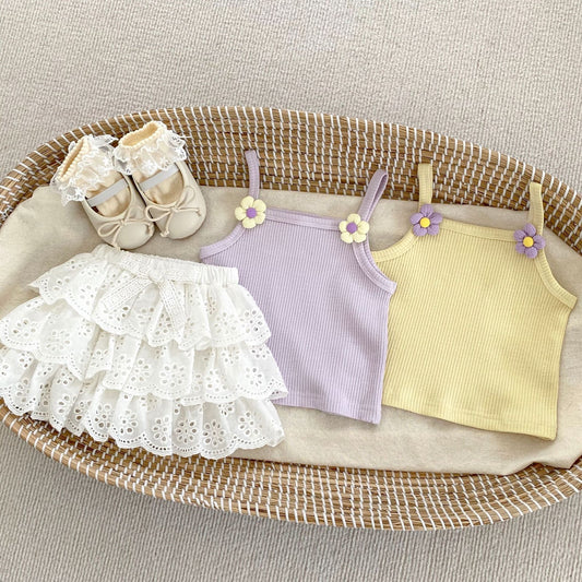 Children's clothing including a white dress, purple tank top, and yellow tank top on a woven basket.